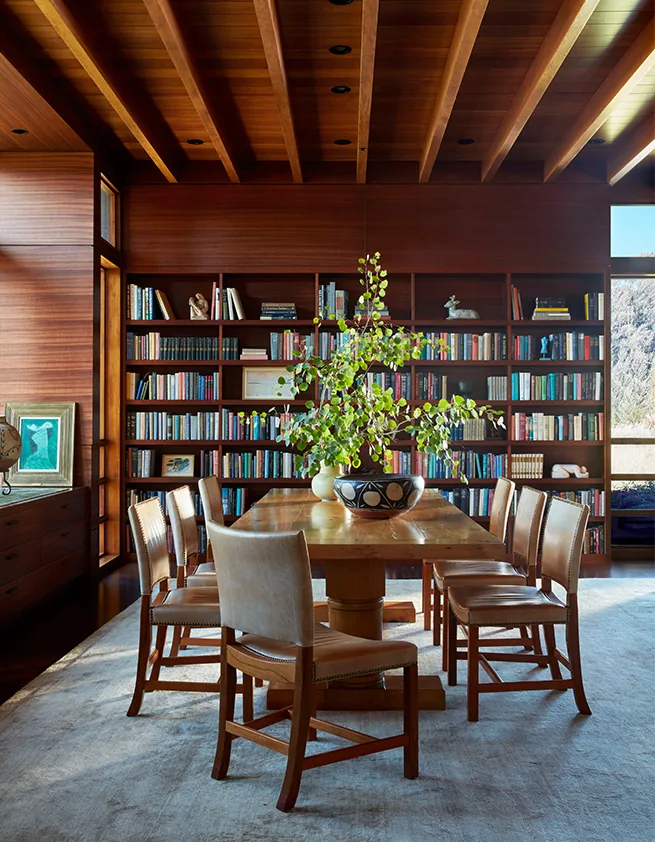 Wood-paneled dining room with a large wooden table and built-in bookcases under an exposed beam ceiling.