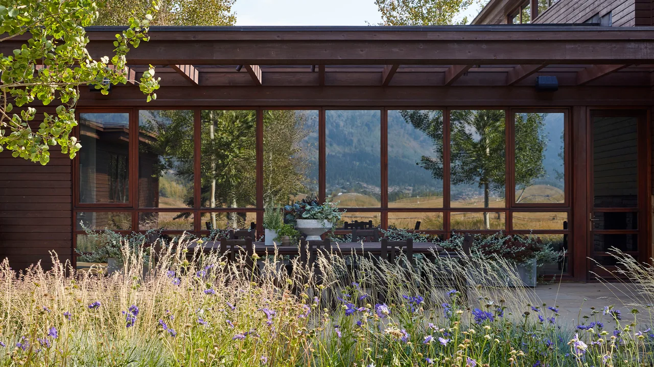 The Home on the Ranch features large wood-framed windows reflecting natural landscape, with wildflowers in the foreground of the outdoor patio area.