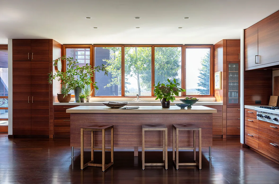 The kitchen features floor-to-ceiling wood-paneled cabinetry, a large island with stools, and expansive wood-framed windows overlooking trees.