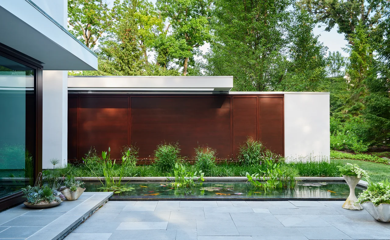 Patio with a rectangular water feature in front of a modern home with dark wood paneled walls and lush greenery.