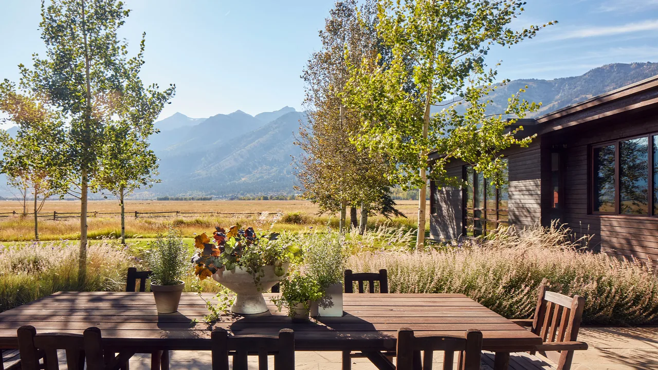 A wooden outdoor dining table and chairs are set on a patio with views of mountains and surrounding greenery.