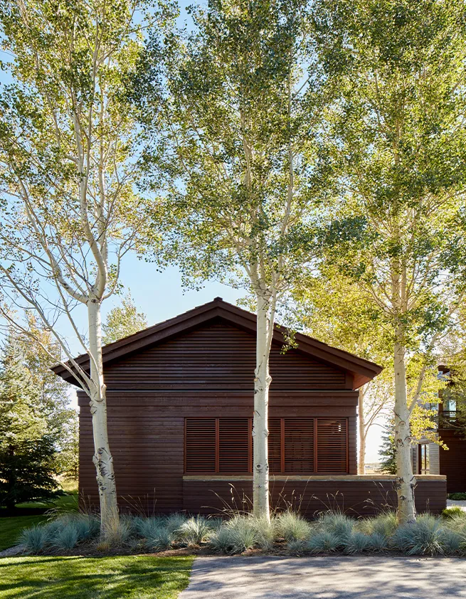 The Home on the Ranch features brown wood siding with a gable roof and is set among mature trees and landscaped grasses.