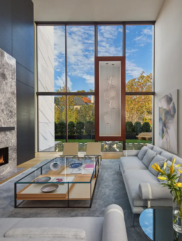 Living room features floor-to-ceiling windows, a fireplace, and light-colored furniture with autumn foliage outside.