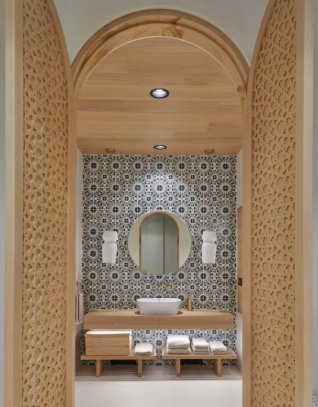 Bathroom interior features a patterned tile accent wall, round mirror, and a floating wooden vanity with towels and decorative sconces.