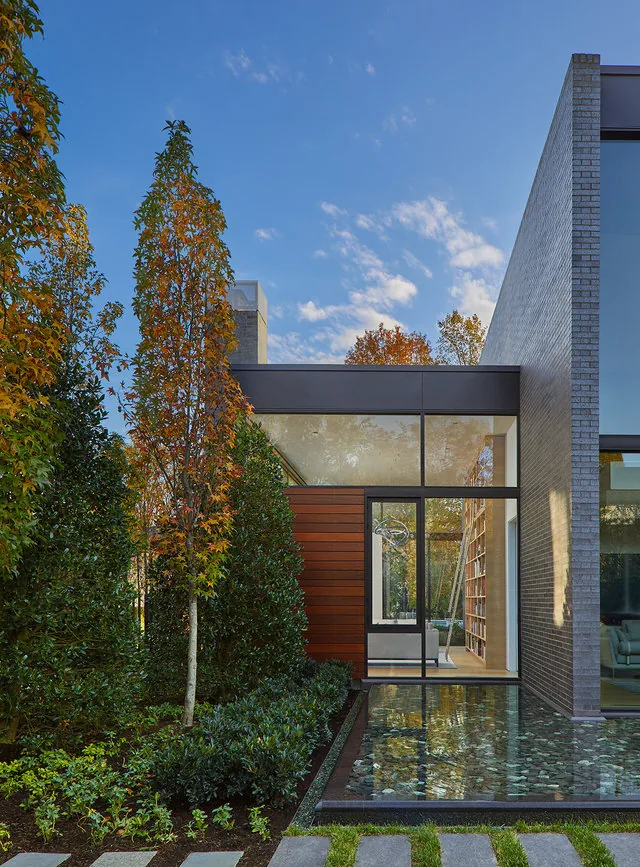 The Urban Woodland House features a mixed material façade with brick, wood siding, and expansive glass walls adjacent to a reflecting pond.