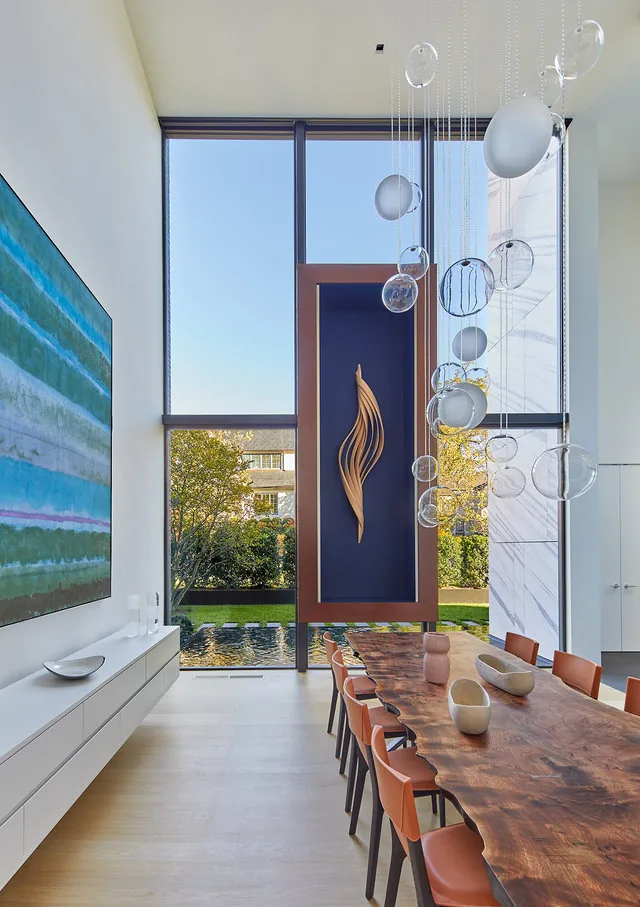 Dining room in the URBAN WOODLAND HOUSE features floor-to-ceiling windows and a live edge wooden dining table with modern lighting.