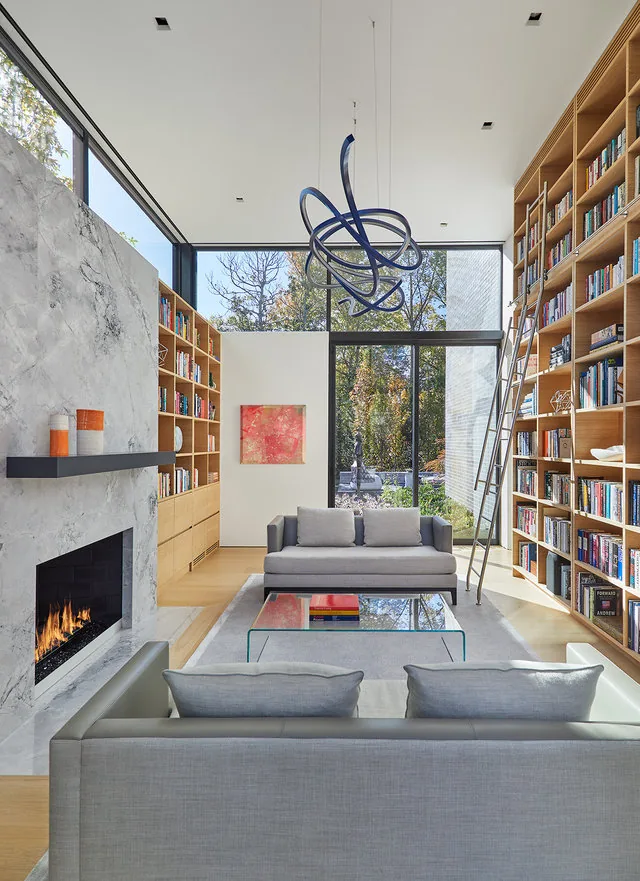 Living room with double-height ceiling, floor-to-ceiling glazing, marble fireplace, and extensive built-in wooden bookshelves.