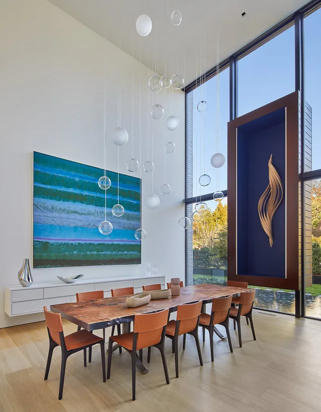 Double-height dining room featuring a wooden table with brown seating, pendant glass globe lights, and expansive floor-to-ceiling windows overlooking autumn trees.