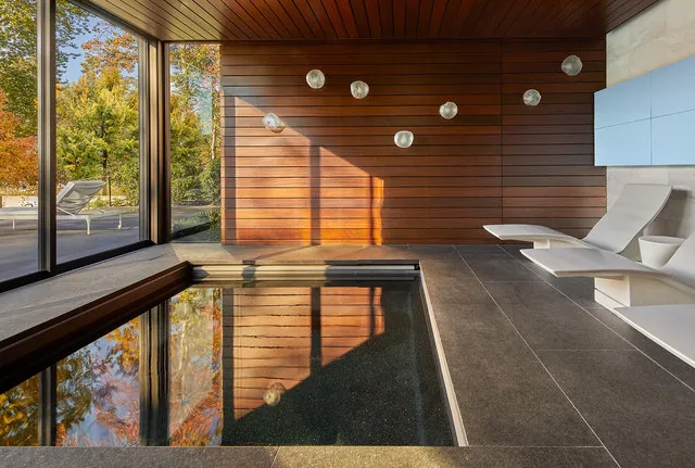 Indoor pool room with floor-to-ceiling glazing, wood wall paneling, and modern lounge chairs.