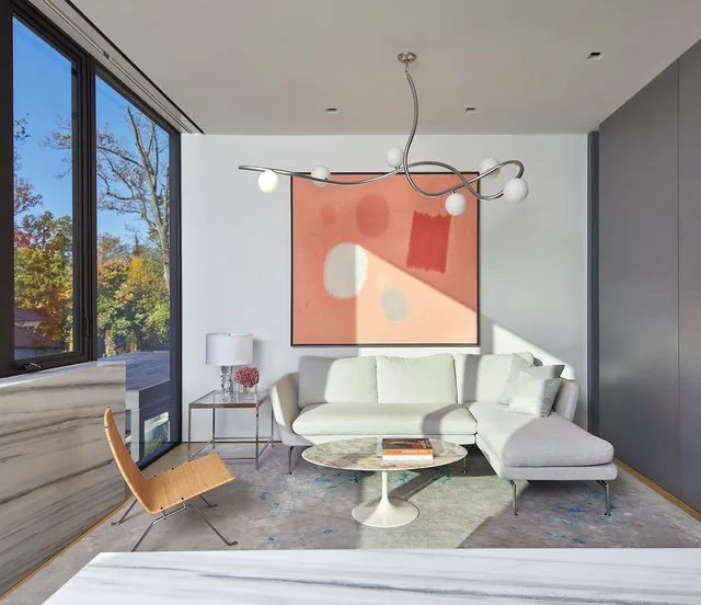 Living room of the Urban Woodland House features a light gray sectional sofa, abstract wall art, and a large window with views of trees.