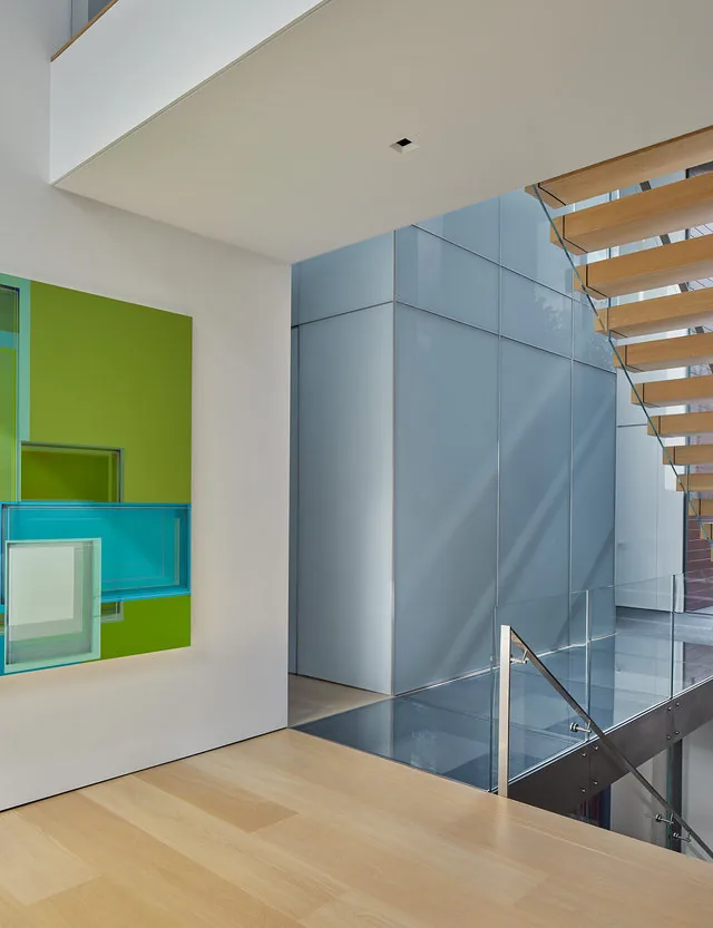 A modern interior features light wood flooring, floating stairs with glass balustrade, and bright geometric wall art.