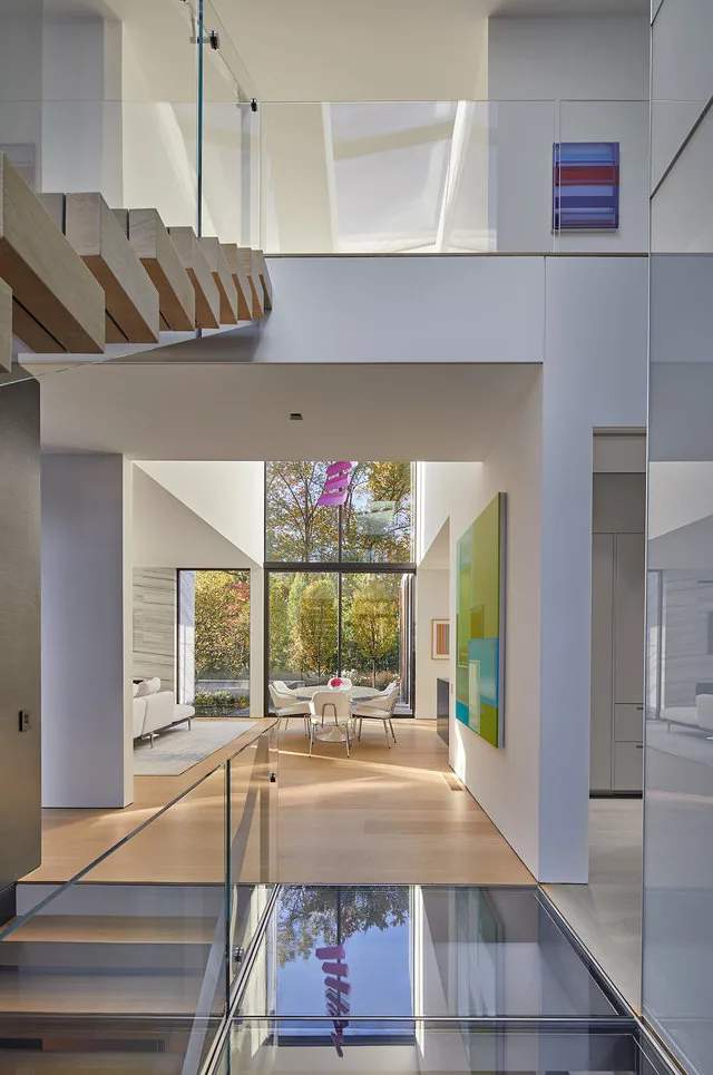 The Urban Woodland House by Robert Gurney Architects features a floating wood staircase, glass railings, and double-height ceiling with large floor-to-ceiling windows.