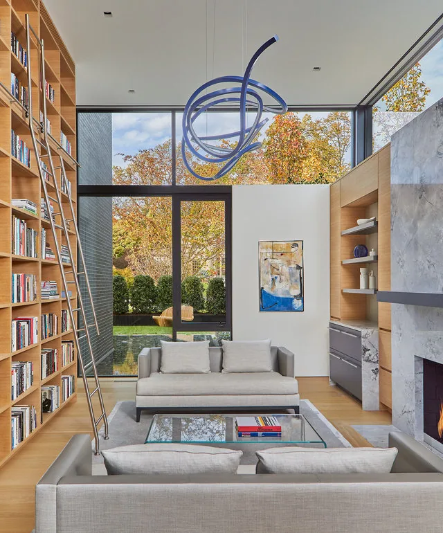 The URBAN WOODLAND HOUSE living room features floor-to-ceiling windows, built-in bookshelves, and a glass coffee table with gray seating.