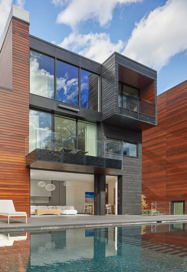Exterior view of a modern house featuring black and wood siding with large glass windows and balconies above a reflective pool.