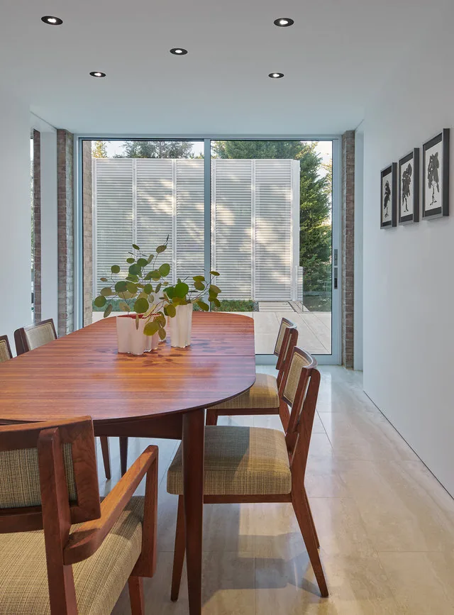 A dining room features a wooden table and chairs with sliding glass doors leading to an outdoor area with privacy panels.