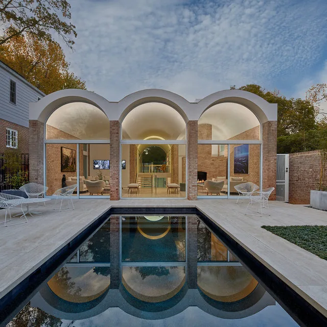 Midcentury modern house featuring brick walls and barrel vault arches, with a rectangular pool and white metal chairs in the patio area.