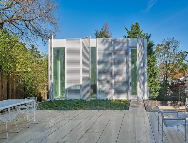 Modern exterior of a small building with white slatted panels and glass doors opening to a tiled patio with metal chairs and a garden area.