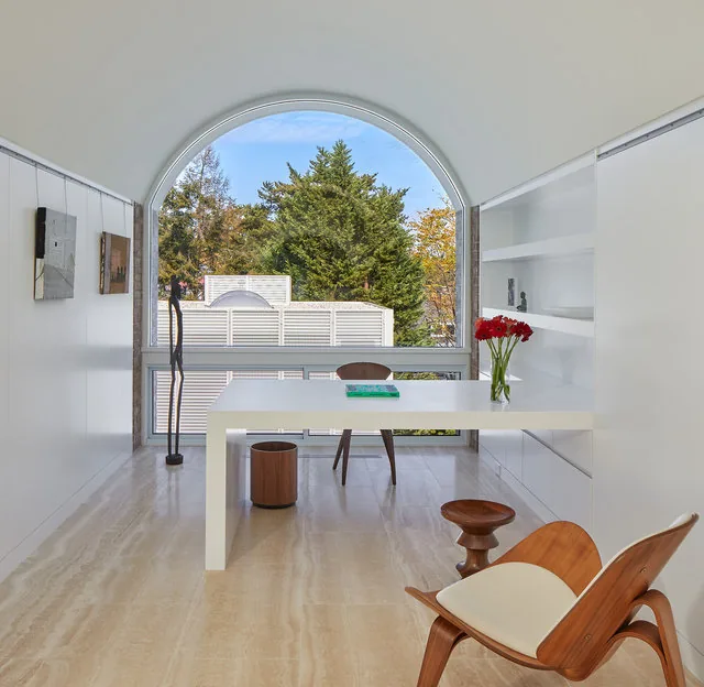 Office space with a large arched window, minimalist white desk, and modern wooden chairs.