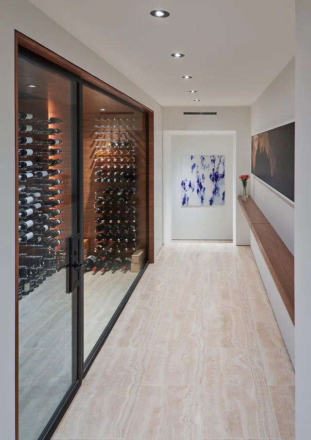 A narrow hallway features recessed ceiling lights, light-colored tile floor, a glass wine storage with wooden back, and wooden wall shelf with flowers.