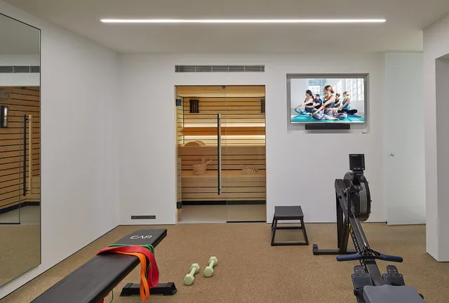 Home gym features rowing machine, exercise bench, wall mirror, and a sauna with glass doors.