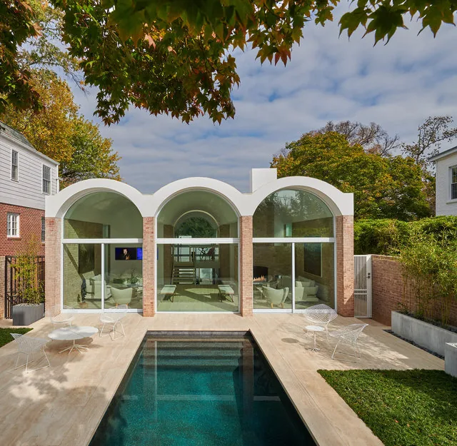 Modern rear addition with three arches and large glass windows overlooking a rectangular pool and patio with white metal furniture.