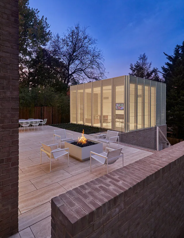 The Slayton House renovation features an illuminated glass and metal addition beside an outdoor firepit patio with white seating.