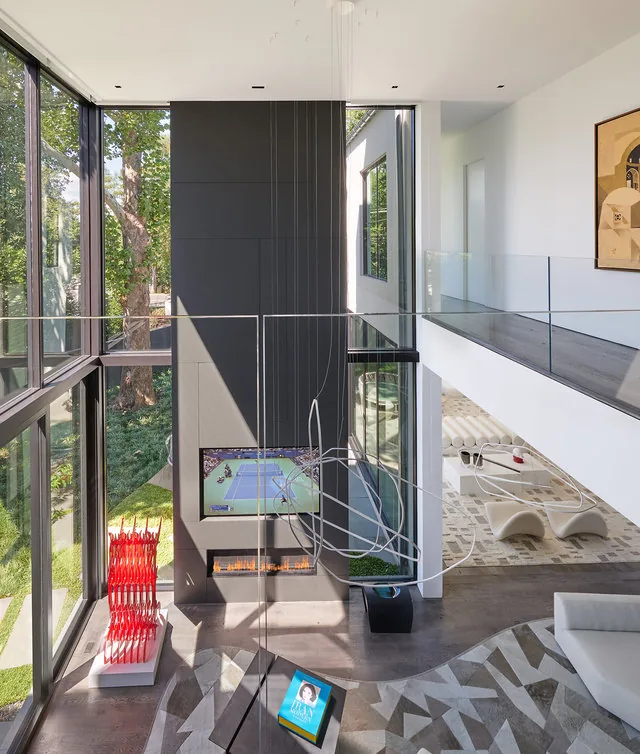 Modern two-story living room with floor-to-ceiling windows, a wall-mounted TV above fireplace, sculptural lighting, and a glass railing on upper level.