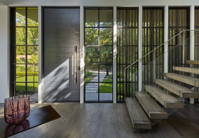 Entry of Phillips Park House featuring a wood-panel door, floor-to-ceiling grid windows, and floating stairs with metal railing.
