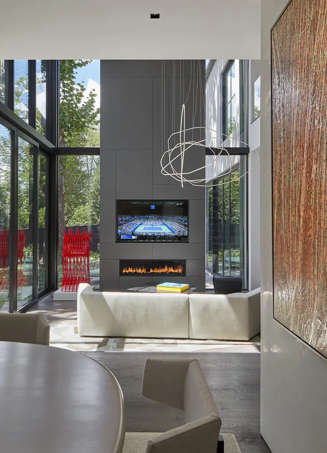 High-ceiling living room features floor-to-ceiling glazing, a linear fireplace, and a modern pendant light above a white sofa.