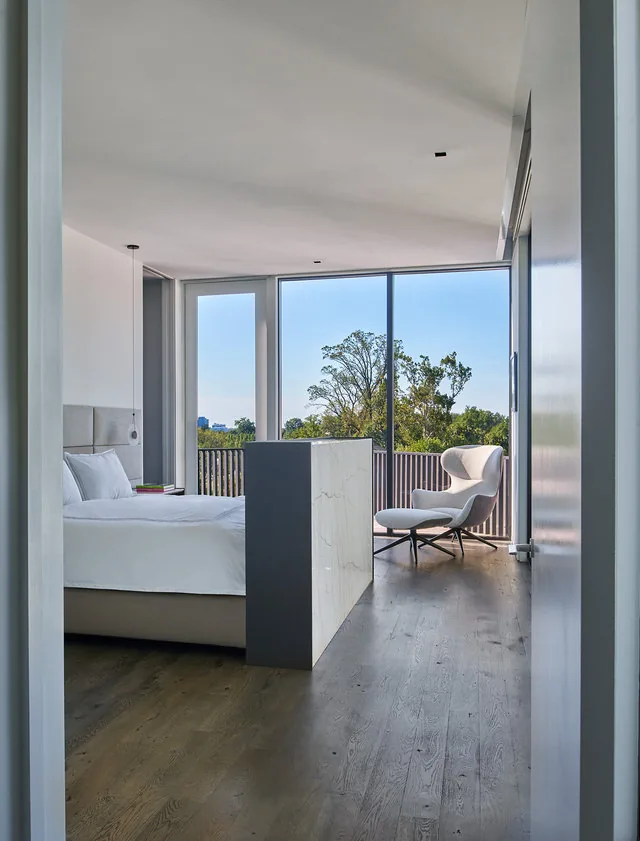 Light-filled modern bedroom featuring floor-to-ceiling glazing and a minimalist lounge chair overlooking trees.