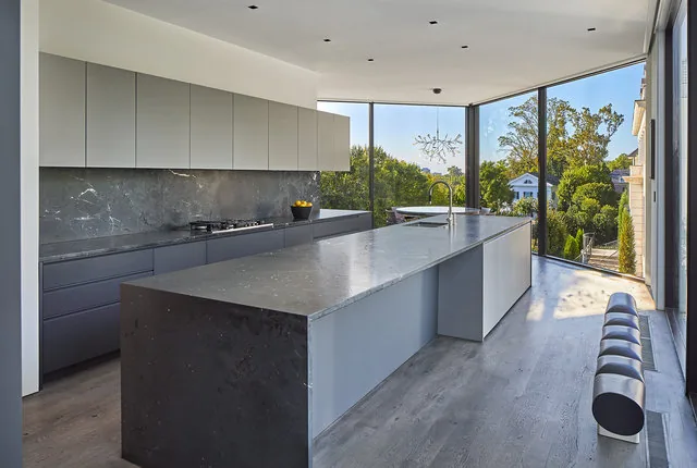 PHILLIPS PARK HOUSE kitchen features a large island with gray stone countertops and corner floor-to-ceiling windows.
