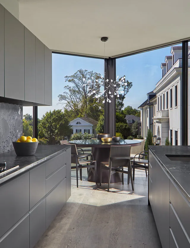 PHILLIPS PARK HOUSE kitchen features gray cabinetry, marble countertops, and a round dining table in a corner with floor-to-ceiling glazing.