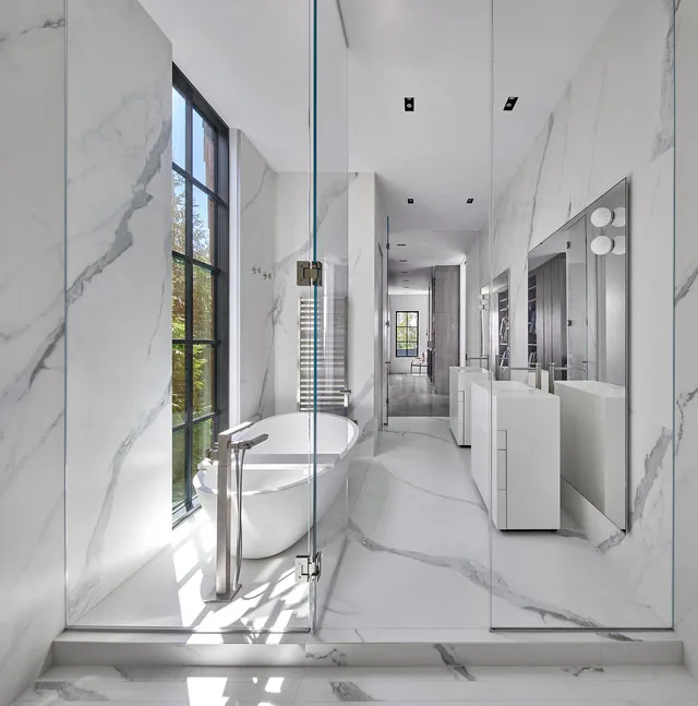 The Phillips Park House bathroom features white marble surfaces, a freestanding tub, and black-framed floor-to-ceiling windows.