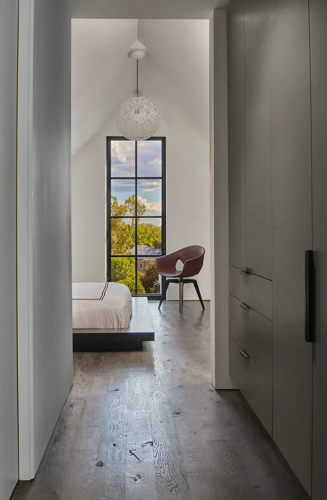 Vaulted hallway views a bedroom with a floor-to-ceiling grid window and a modern chair near the bed.