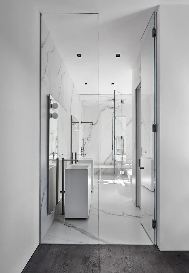 The Phillips Park House bathroom features white marble surfaces, dual pedestal sinks, and a frameless glass shower.