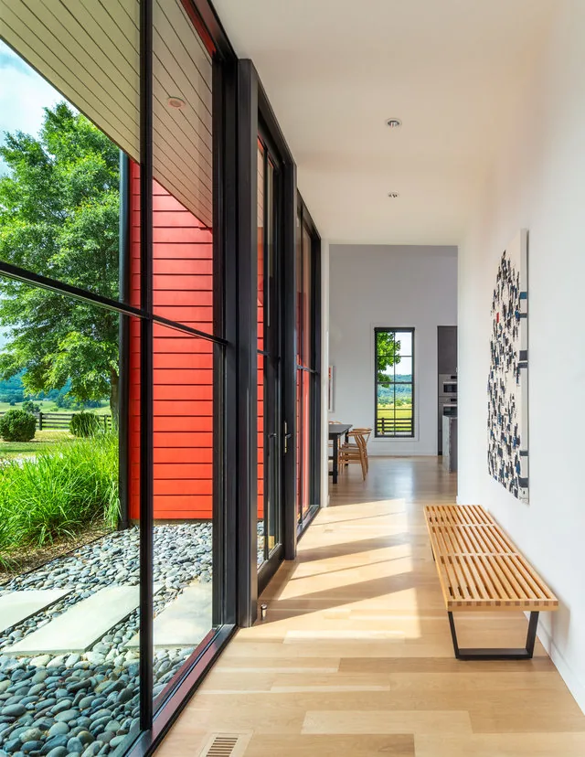 Hallway with floor-to-ceiling windows and light wood bench in a house featuring red exterior siding.