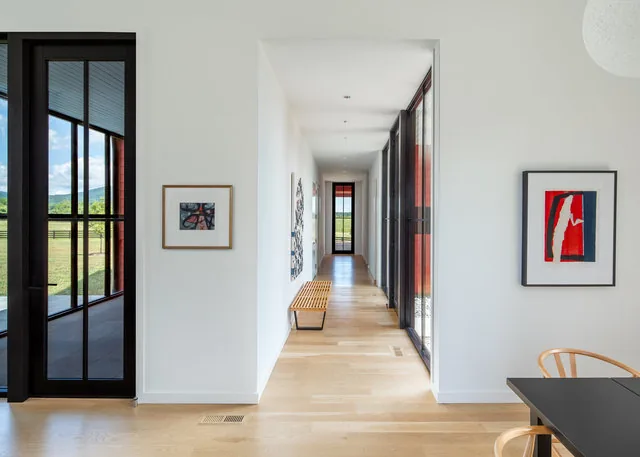 Hallway in Etlan Farm House features floor-to-ceiling black-framed glass doors, light wood flooring, and minimalist artwork.