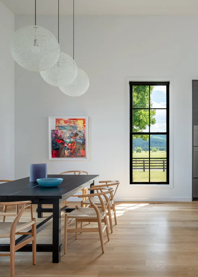 Dining room with black table, natural wood chairs, pendant lights, and a window framing a rural pasture view.