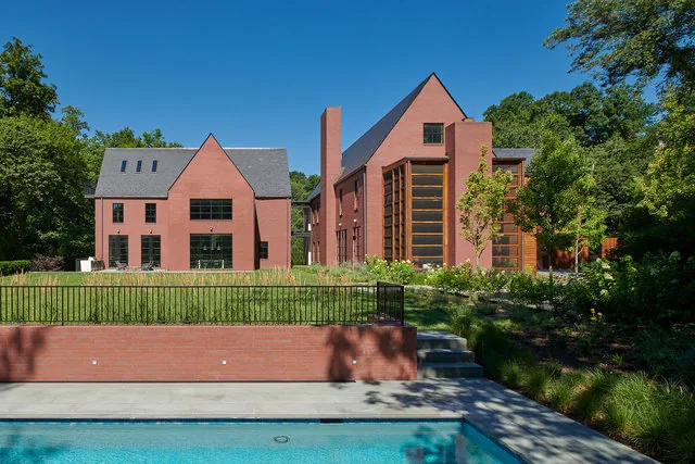The 30TH STREET HOUSE by Robert Gurney Architects features steep gable roofs and a symmetrical red brick facade with a swimming pool and iron fence in front.