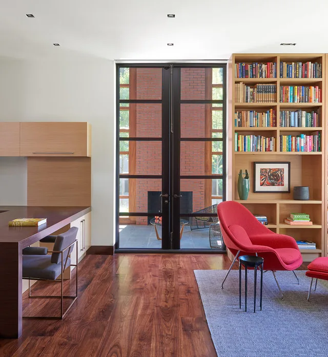 Living area with built-in wood bookshelves, red lounge chair, and floor-to-ceiling glass doors opening to a covered patio with brick walls.