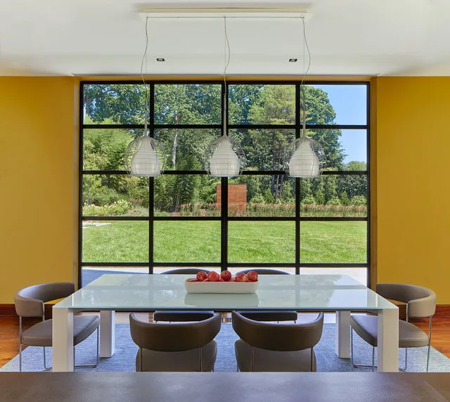 Dining area features a large grid window with black frames, a white table, six brown chairs, and three pendant lights.