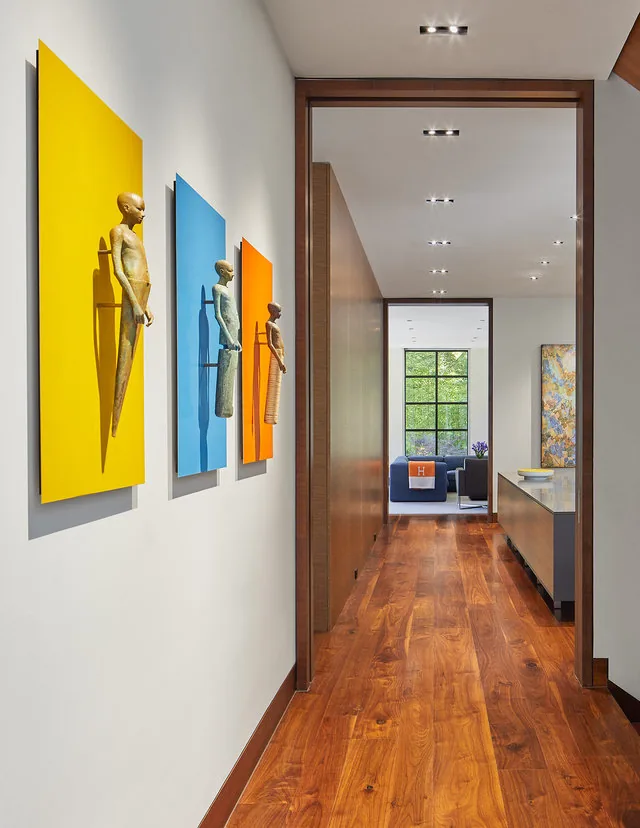 A hallway with wood flooring and colorful art panels leads to a living room featuring a large black-framed picture window.
