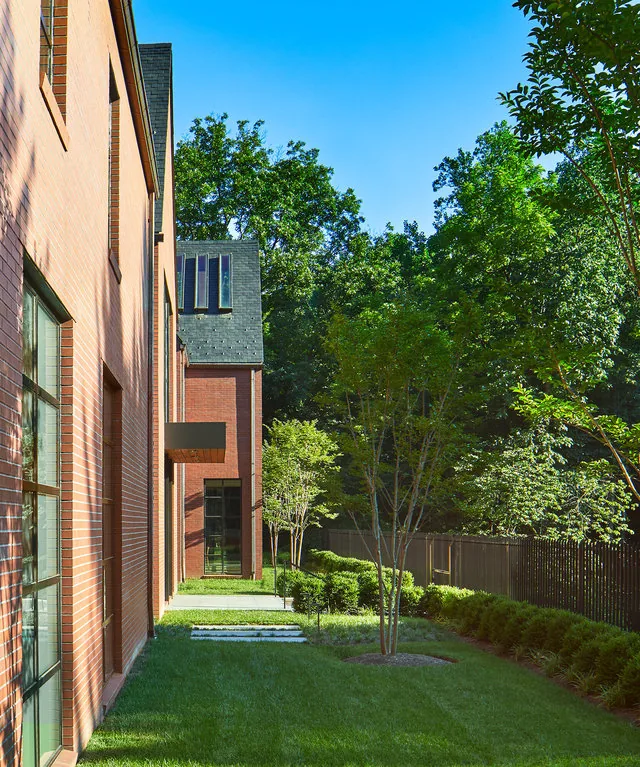 Brick house side view with symmetrical large rectangular windows, landscaped garden, and iron fence in a wooded area.