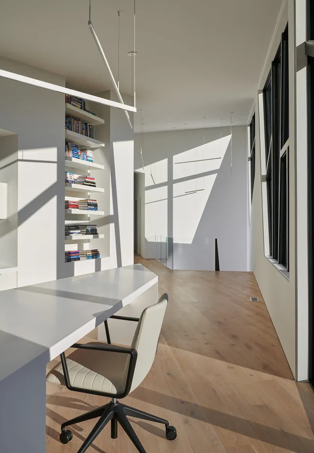 PTR HOUSE interior hallway with built-in bookshelves, large windows, and minimalist white desk and office chair.