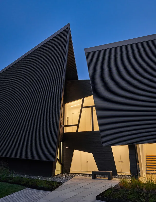 PTR House by Robert Gurney Architects features angular dark ribbed siding and triangular windows with illuminated interiors at dusk.