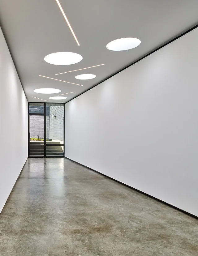 Hallway with polished concrete floor, white walls, circular skylights, and linear ceiling lights in PTR HOUSE.