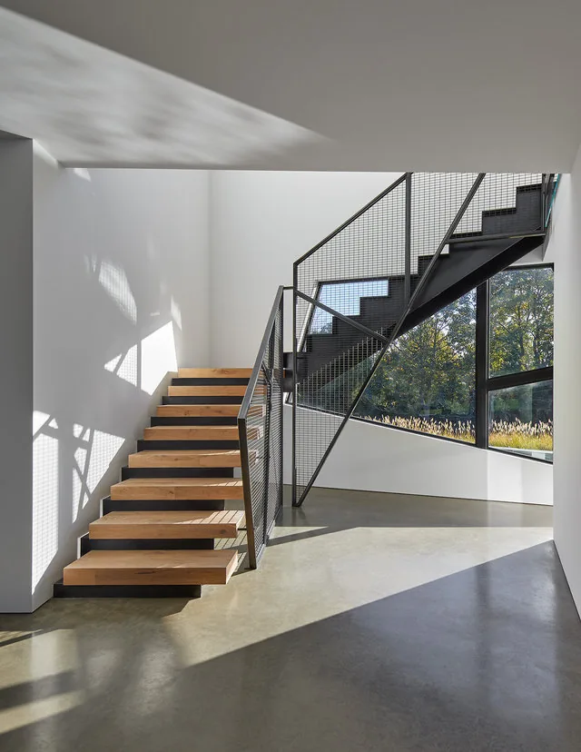 Modern angular staircase with floating wood steps and mesh metal railing in a brightly lit space at PTR HOUSE.