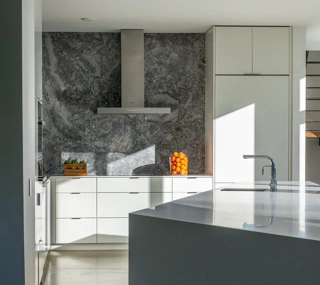 Modern kitchen featuring white cabinets, a polished stone countertop island, and a dark textured marble backsplash with a stainless steel vent hood.