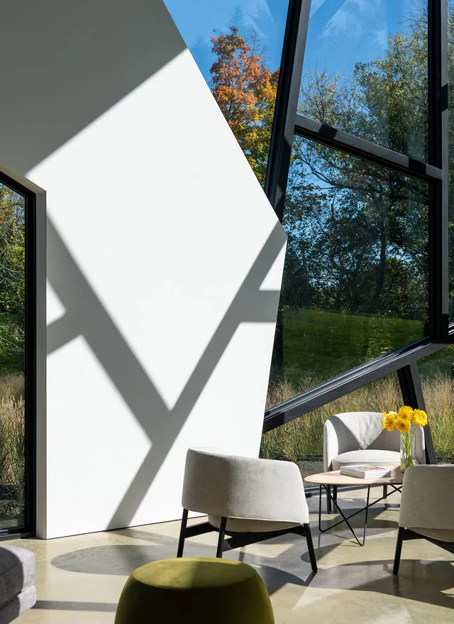 Sunlit seating nook with light gray armchairs and a modern coffee table beside angular floor-to-ceiling windows.