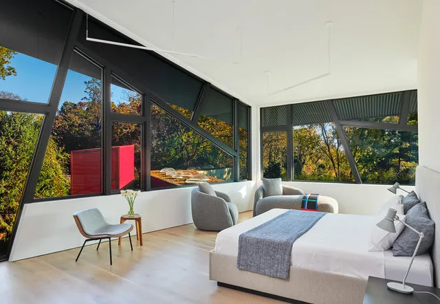 Bedroom interior showcasing angled large windows with black frames and an autumn forest view.